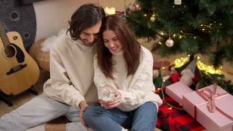 Happy-couple,-a-brunette-guy-in-a-white-sweatshirt-and-a-brunette-girl-in-a-White-sweater-who-shakes-the-New-Year's-Sphere-and-looks-at-the-flying-snow-near-pink-gifts-of-a-red-blanket-and-a-green-Christmas-tree