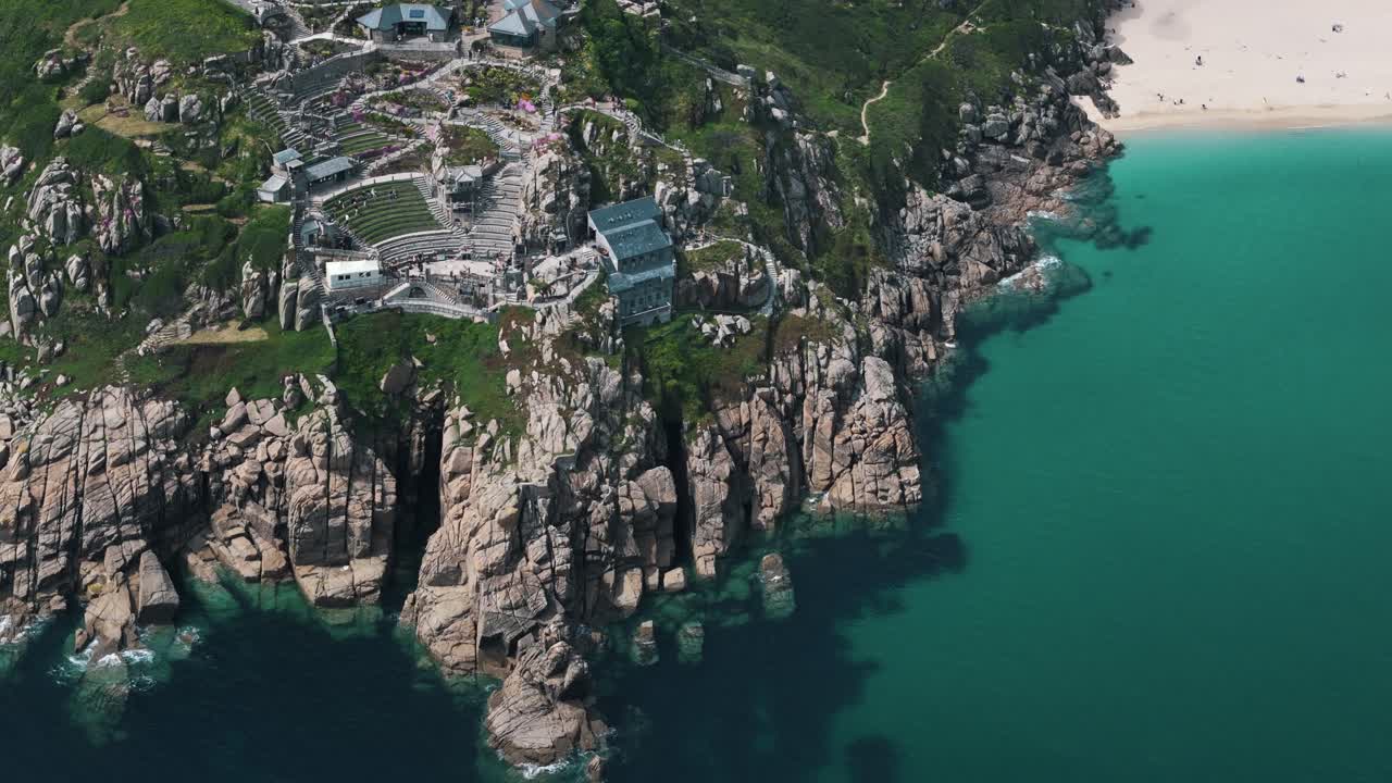 Drone tracks left over Minack Theatres cliffside terraces with blue ocean beyond, aerial establishing overview
