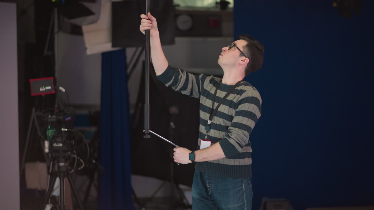 Technician adjusts stage lighting equipment in a studio with a boom pole. Focus on equipment hanging from ceiling while technician operates it for a production setup in broadcast environment