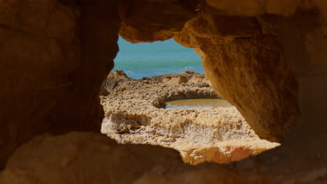 vista de las olas del mar a través del agujero en los acantilados bajos de ocre en praia do evaristo en el algarve, portugal