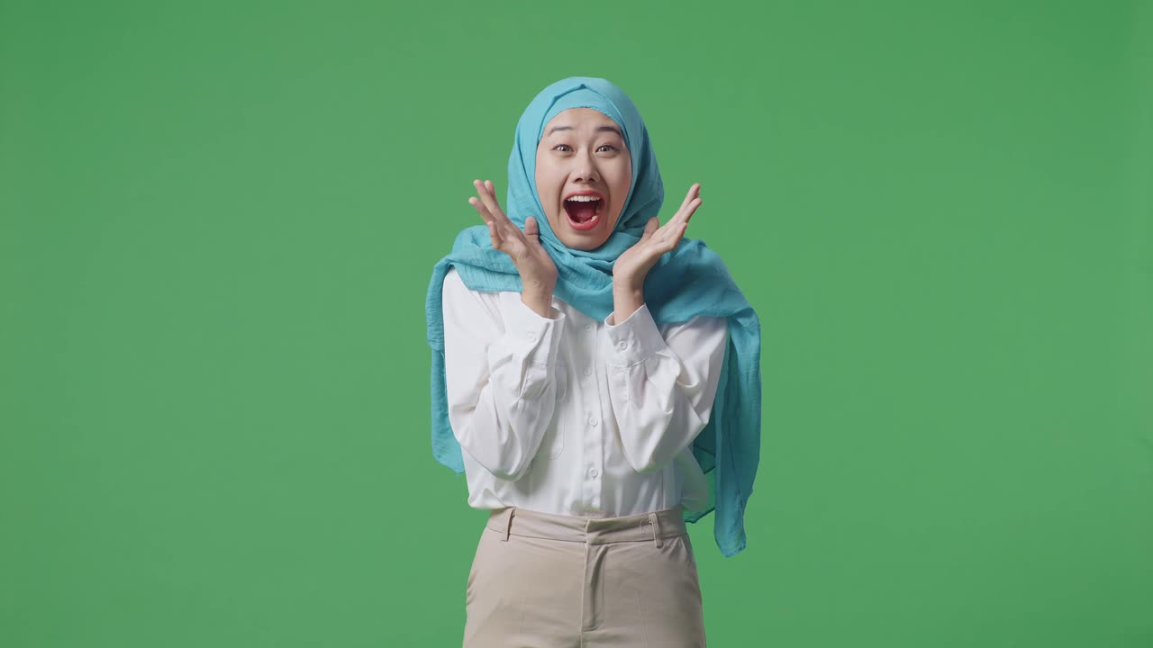 아시아의 무슬림 여성이 미소 짓고 와우라고 말하면서 녹색 스크린 배경 스튜디오에 서 있습니다.