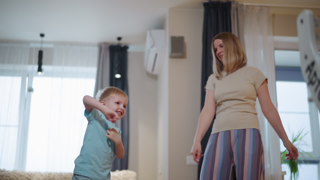 Mother swinging young son playfully by hands in bright living room, child lifted off ground with joy, expressing fun, laughter, excitement, and warmth during family bonding moment in cozy indoor home