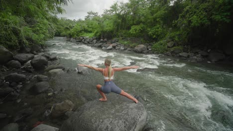 blond woman practicing warrior pose on rock beside jungle river, yoga