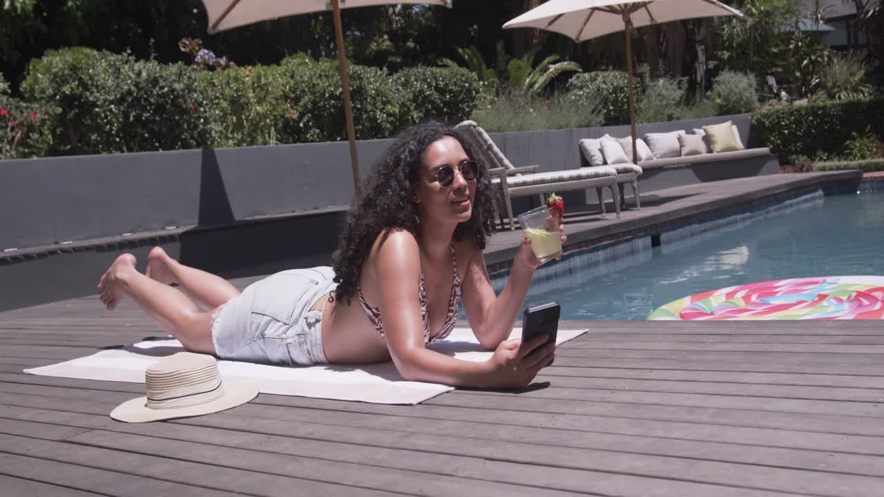 African American woman checking phone, reacting to off-camera call, sipping cocktail poolside
