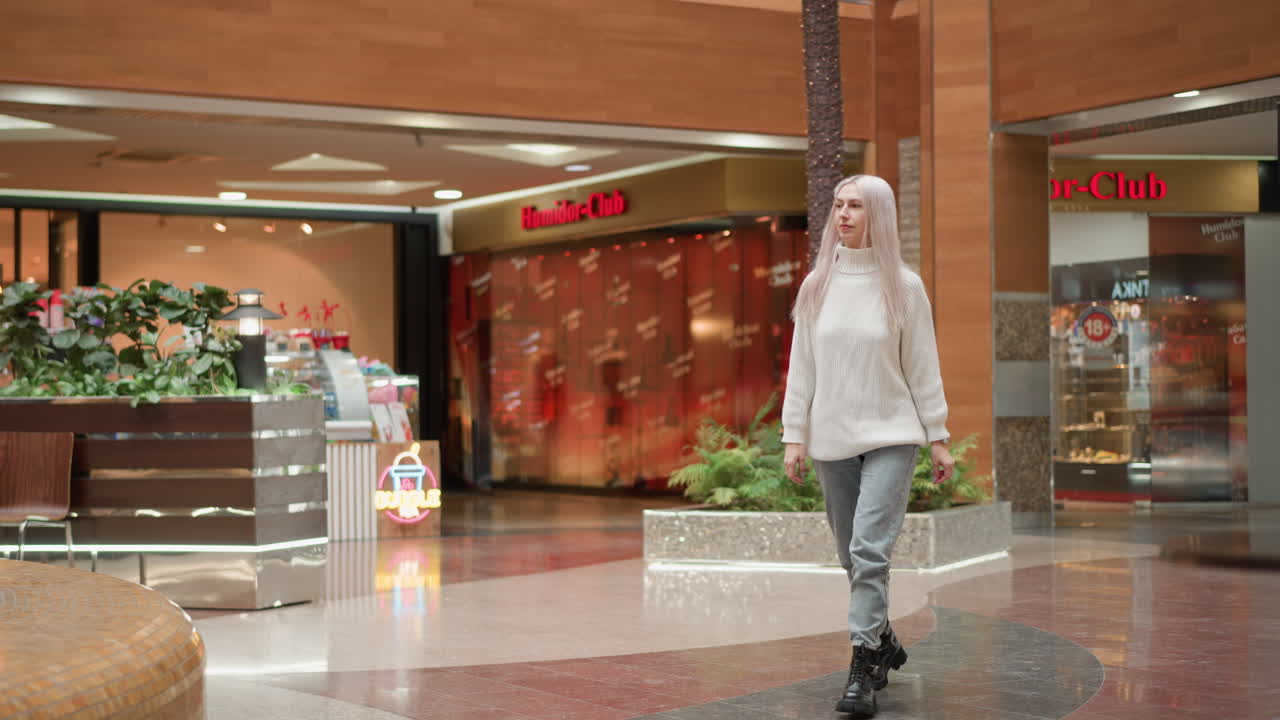 Full length view of modern woman walking through spacious mall interior in knit sweater and jeans under bright ceiling lights with store fronts plants and glass railings blurred backdrop