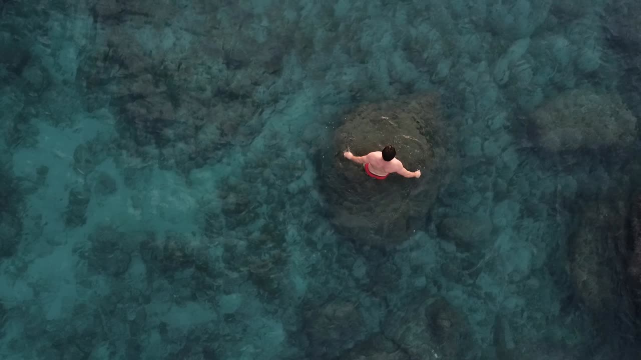 청년은 맑은 청록색 물에서 수영하고, 바로 위의 공중 드론