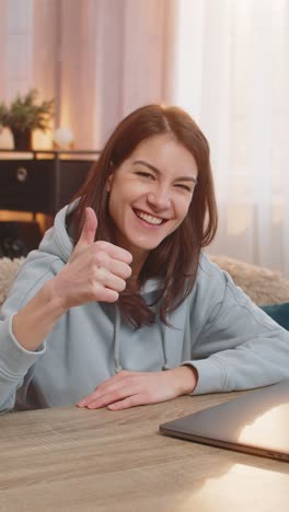 smiling young woman freelancer closing laptop after work and showing thumbs up at table at home