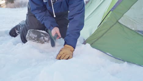 camper hammering the stake on corner of camping tent at winter with an axe then cover with a rock