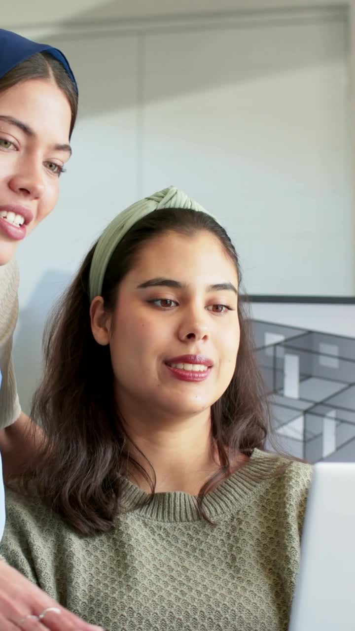 Video verticale: Donna con hijab blu che si china e indica un laptop in un ufficio luminoso, mentre istruisce una collega.