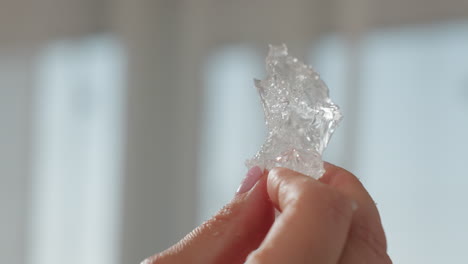 close up of hand with pink nails gently shaking transparent rubber-like soluble substance while bright natural light filters through sheer curtains in soft focus