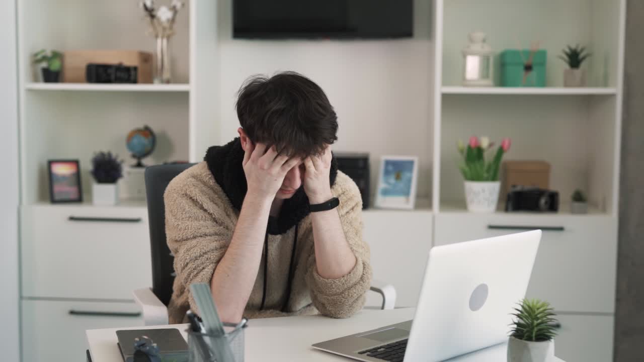 The young man is focusedly working behind his laptop. He indicates that he has a headache. The man is tired from work, and he massages his temples with his hands.