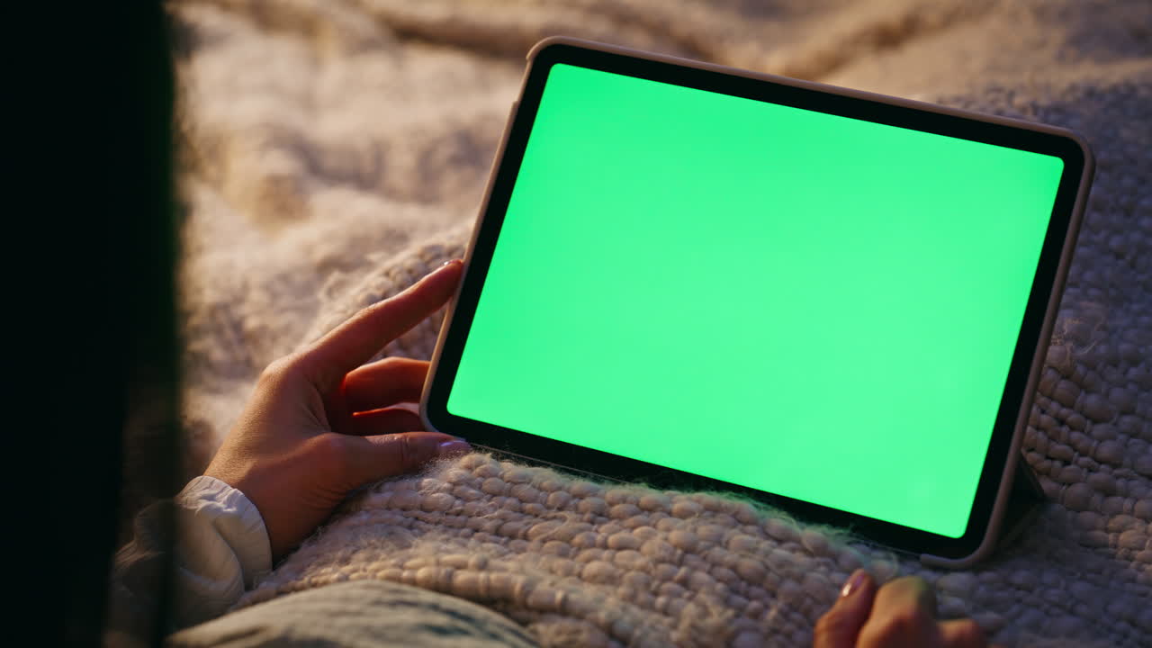 Donna sconosciuta scorre un tablet mockup sdraiata su un letto accogliente, primo piano. Mano che tocca il verde.