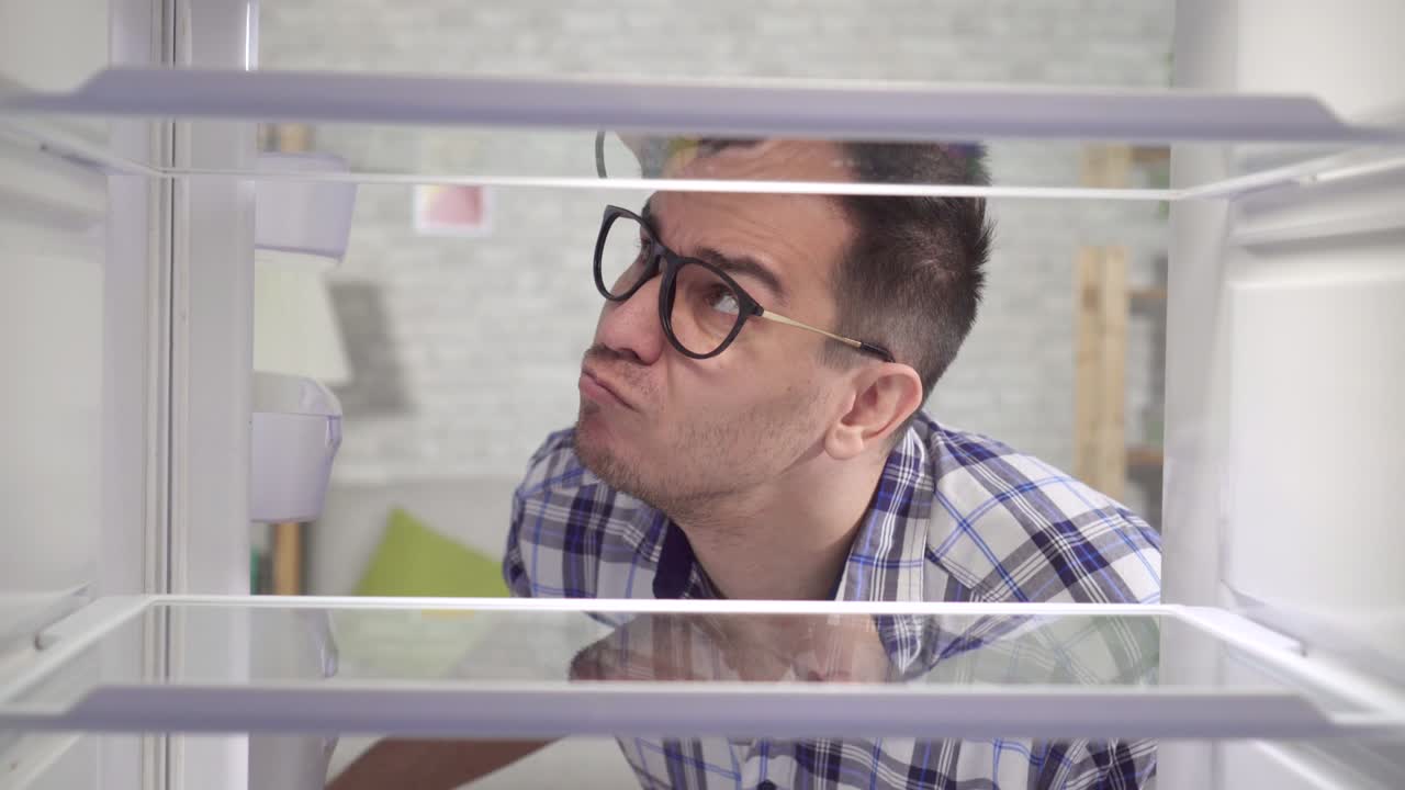 joven pensativo con gafas y una camisa mira en el refrigerador vacío