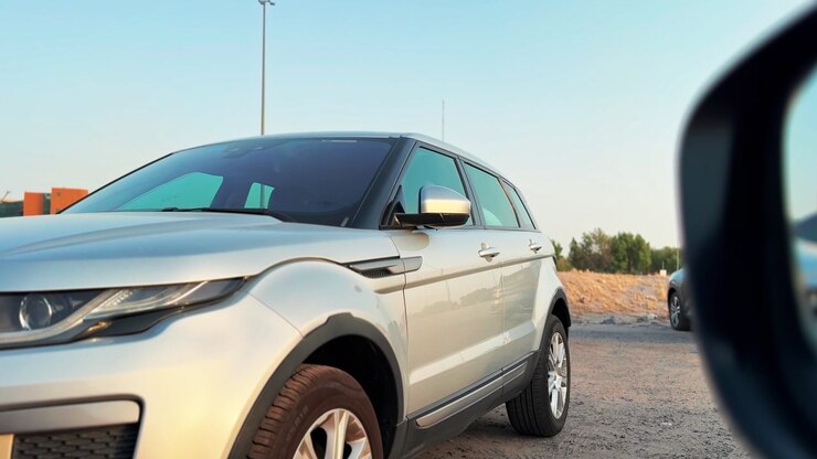 Un SUV argenté est garé sur un terrain en terre poussiéreux en fin d'après-midi. Le véhicule est vu de la perspective d'une autre voiture, avec son rétroviseur latéral visible au premier plan à droite.