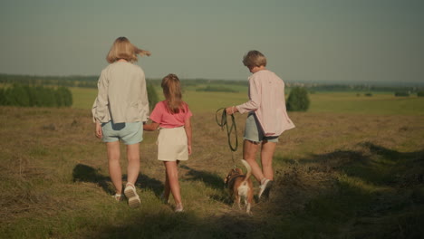 back view of woman walking with dog on leash and mother holding her daughter s hand, strolling through farmland with lush greenery and distant trees under clear blue sky