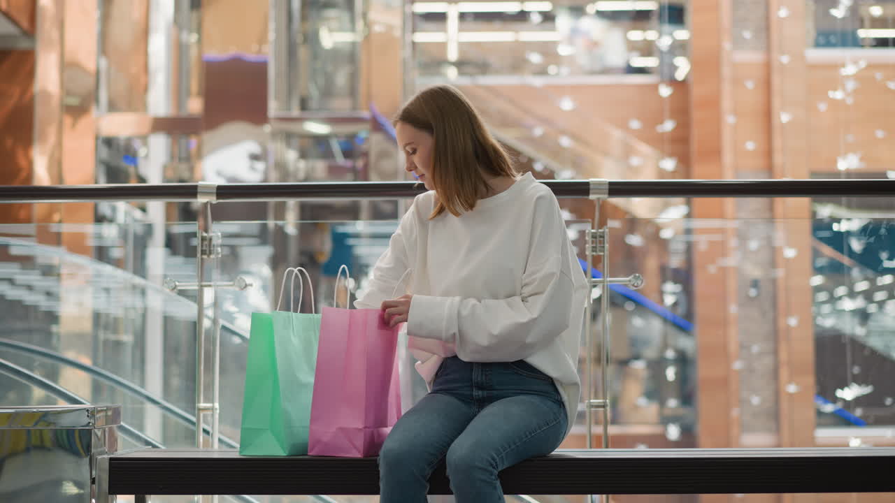 una mujer joven sentada en un banco dentro de un centro comercial moderno recupera un libro de una bolsa de compras rosada, rodeada de un ambiente comercial colorido, barandillas de vidrio y una vista borrosa de las escaleras mecánicas