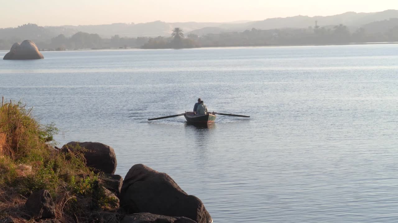 dos hombres remando en un bote en un lago durante el amanecer con rocas en el fondo y en primer plano en asuán, egipto