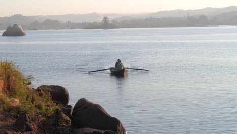 Dos-Hombres-Remando-En-Un-Bote-En-Un-Lago-Durante-El-Amanecer-Con-Rocas-En-El-Fondo-Y-En-Primer-Plano-En-Aswan,-Egipto