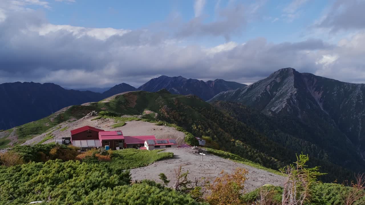 Wide shot of Chogatake Hut perched on a ridgeline with the steep Hotaka mountain range rising behind it under dramatic clouds