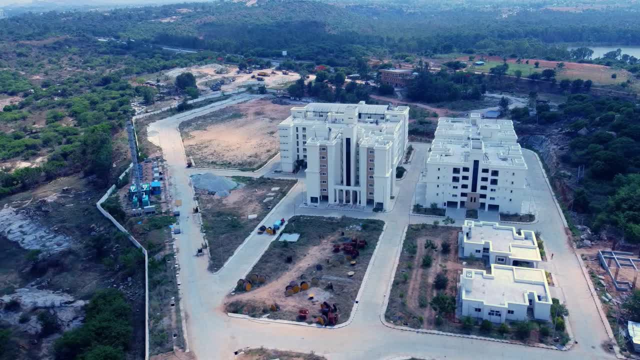 Sweeping aerial view of a large real estate development in India. Modern residential buildings and ongoing construction sites set in a rugged, hilly landscape near a lake, showcasing massive growth
