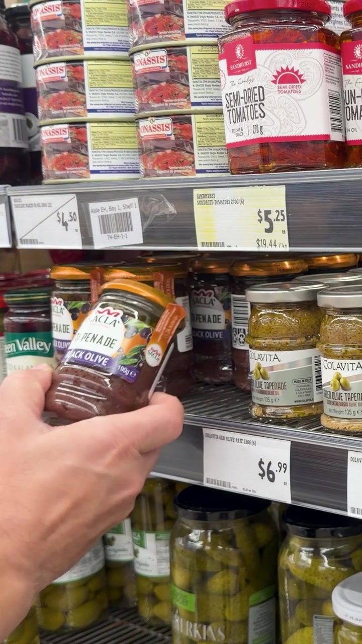 A mão de uma pessoa alcança uma prateleira de supermercado para selecionar e inspecionar um pote de vidro.
