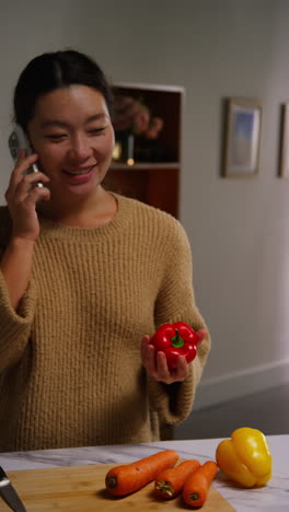 vertical video of woman talking on mobile phone at home preparing fresh vegetables for meal on counter in kitchen