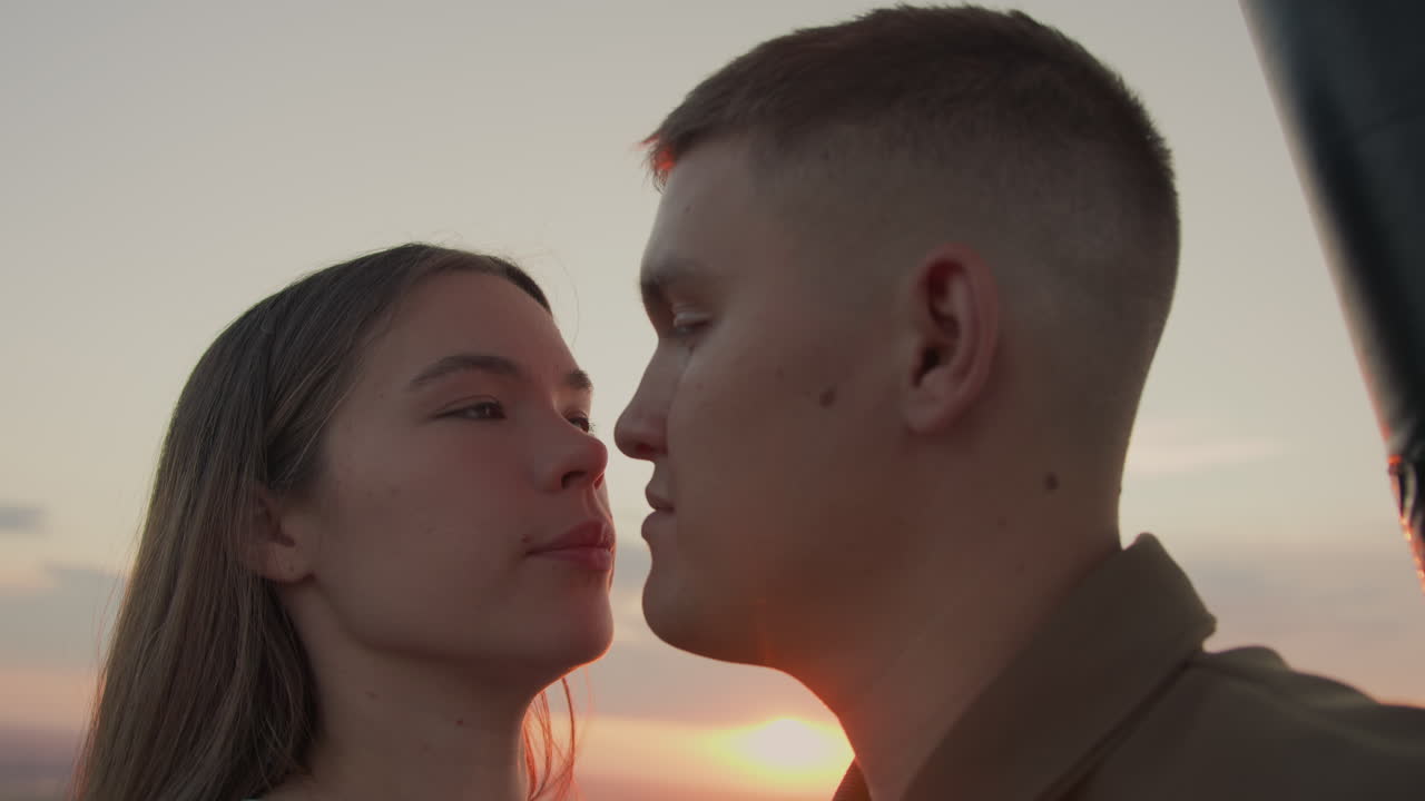 close up couple faces turned toward glowing horizon as balloon basket supports frame silhouette of appreciation and intimacy under pastel sky with distant field panorama at golden hour
