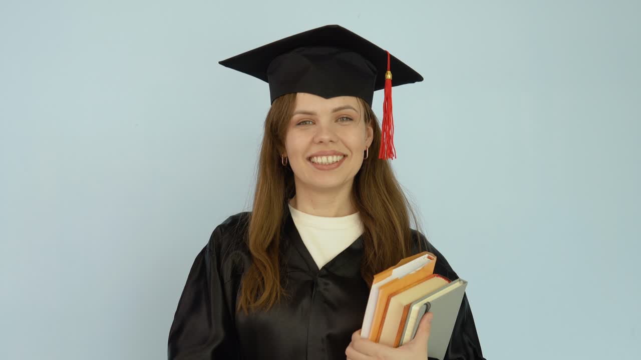 una joven caucásica con una túnica negra y un sombrero de maestro se mantiene erguida sosteniendo libros de texto con una mano y mira a la cámara. retrato de media altura sobre fondo blanco