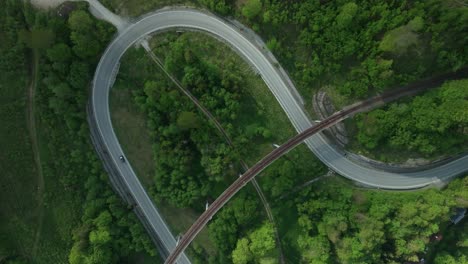 static aerial overhead footage of one car driving on a winding road intersected by an old railway bridge in the middle of a forest