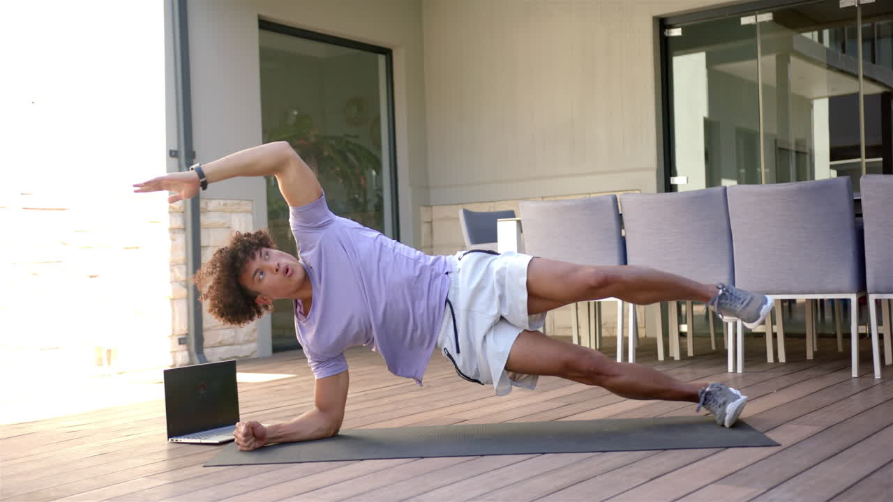 Exercising on yoga mat, man doing side plank with laptop on patio