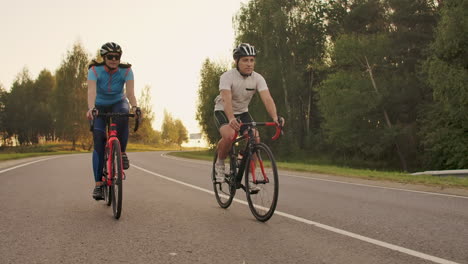 Dos-Ciclistas,-Un-Hombre-Y-Una-Mujer,-Viajan-Por-La-Carretera-En-Bicicletas-De-Carretera-Con-Cascos-Y-Ropa-Deportiva-Al-Atardecer-En-Cámara-Lenta