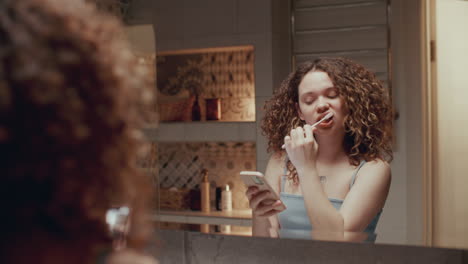 chica cepillándose los dientes y usando el teléfono frente al espejo del baño
