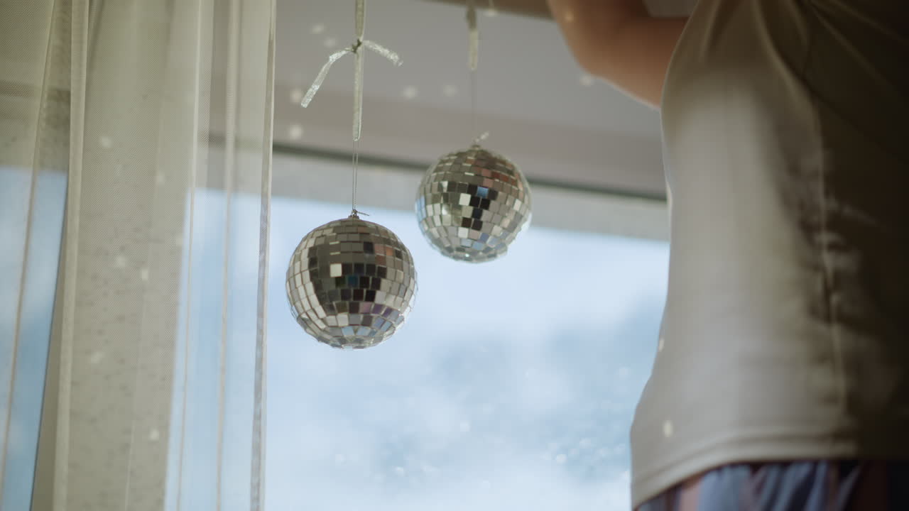 Woman adjusting shiny disco balls hanging near window with sheer curtains, sunlight reflecting on mirrored surfaces creating sparkling festive atmosphere and bright decorative detail
