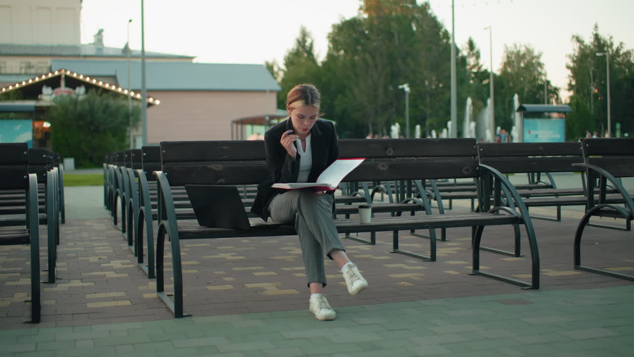 Focused student seated outdoor on wooden bench reading book with laptop beside her and coffee cup in urban setting surrounded by empty benches, paved ground, trees, and soft evening light