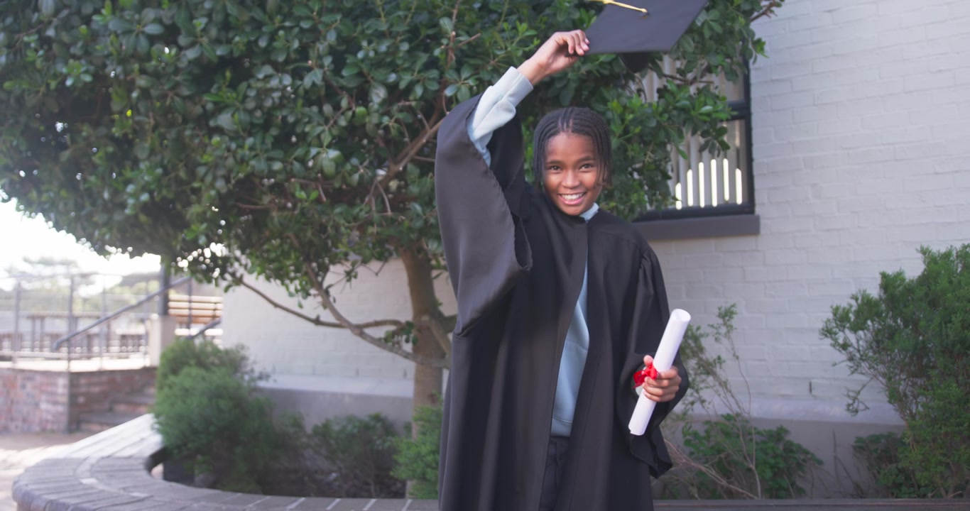 Receiving ribboned diploma African American teen male in black gown tossing cap skyward on campus