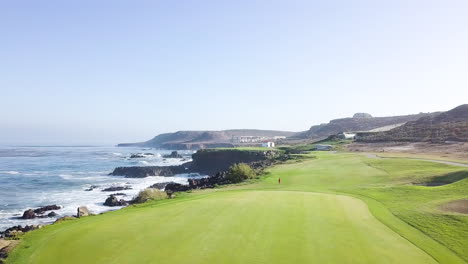 drone flys low over green with a slight right pan with views of a waves crashing on rocks at a beach side golf course and baja's desert landscape