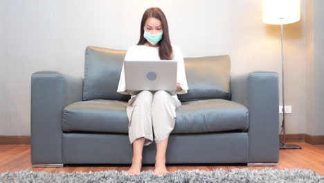 Asiatische-Frau-Mit-Gesichtsmaske,-Die-Auf-Dem-Sofa-Sitzt-Und-Auf-Laptop-Computer-Tippt,-Vollbild