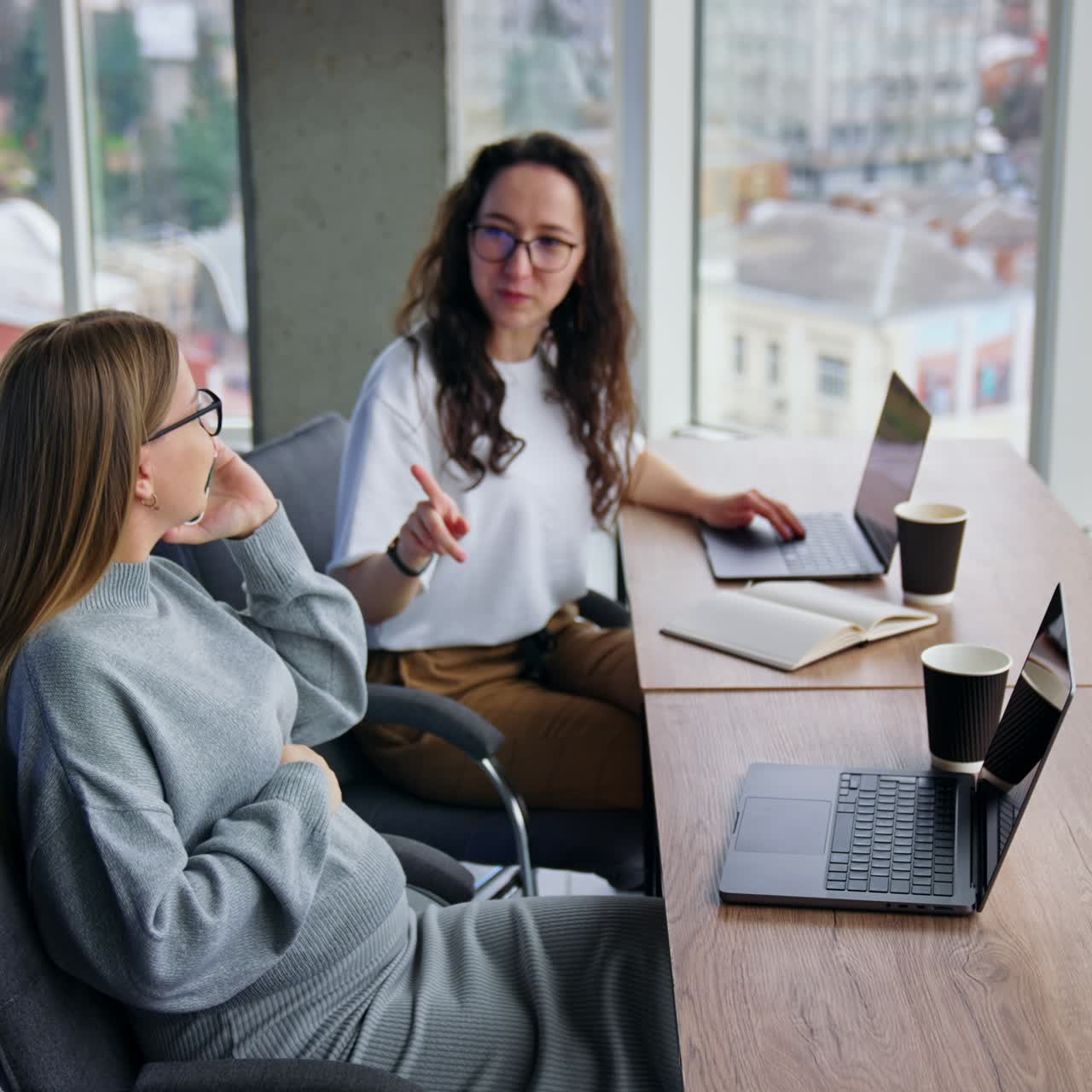 Giovani donne che lavorano in un ufficio moderno. Una donna bionda incinta parla al telefono. Una collega mora lavora al suo laptop. Finestre panoramiche con paesaggio urbano sullo sfondo.