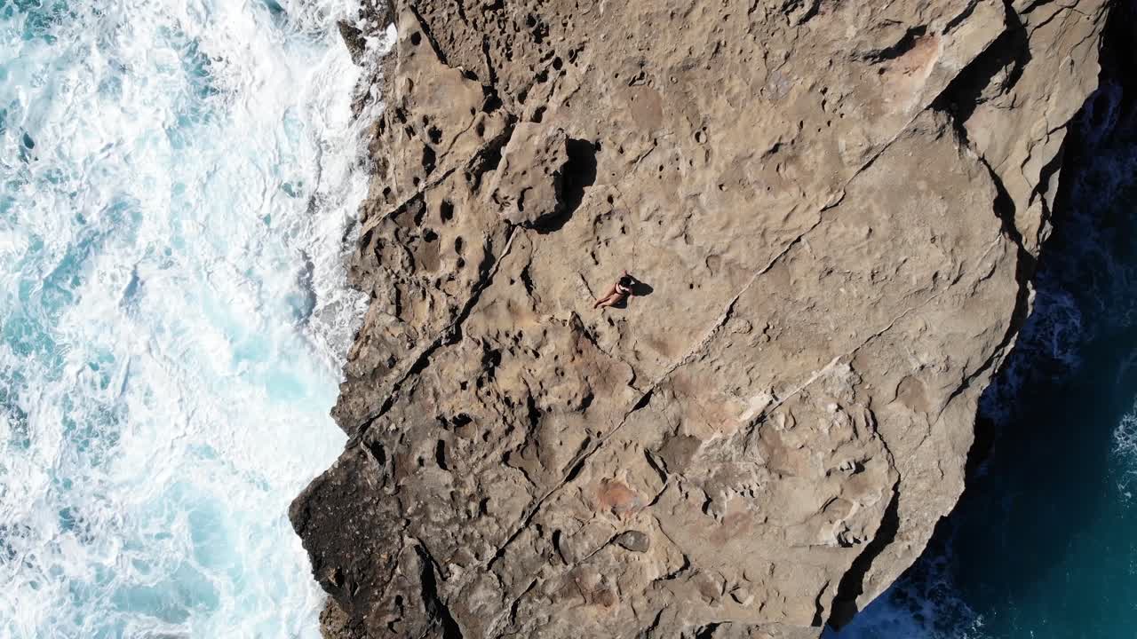 Aerial, top down, drone shot of a woman laying on a cliff, while waves hit the coast of Mallorca, sunny day, in Spain
