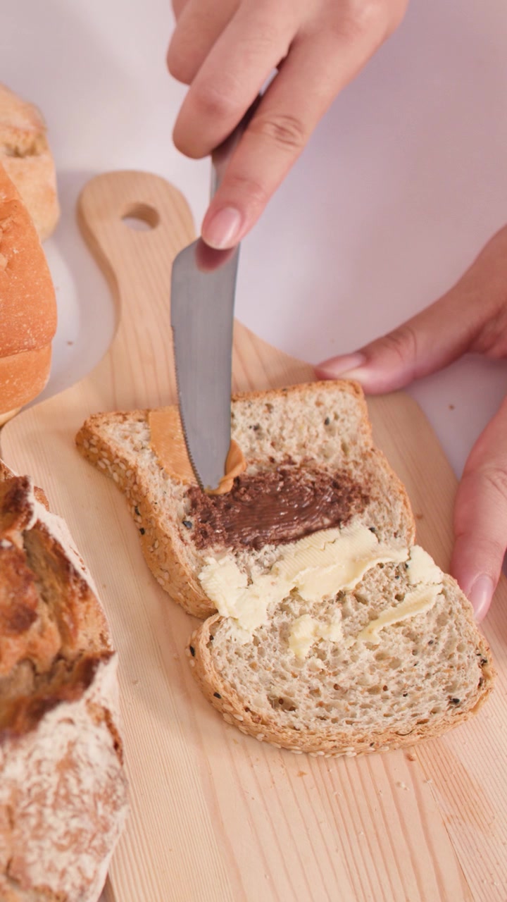 Close-up of hands spreading peanut butter on multigrain bread surrounded by fresh sourdough and baguettes.