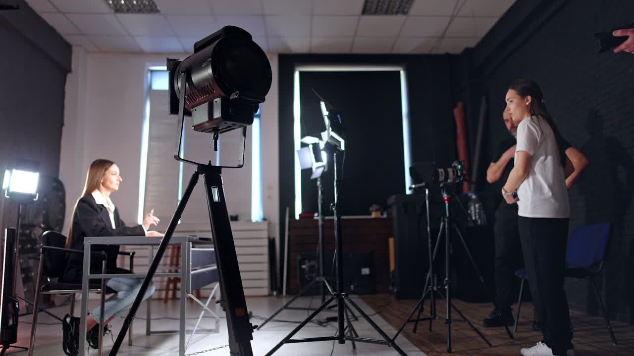 Professional photo studio with all the necessary equipment. Blogger lady sits at desk talking and girl standing opposite taking video on her smartphone.