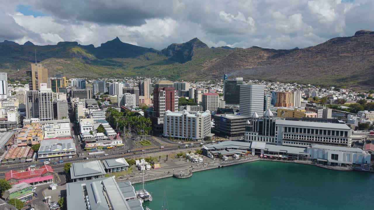 Aerial drone shot capturing the modern Port Louis skyline and the Caudan Waterfront set against the dramatic, rugged Moka Mountains of Mauritius. City meets nature
