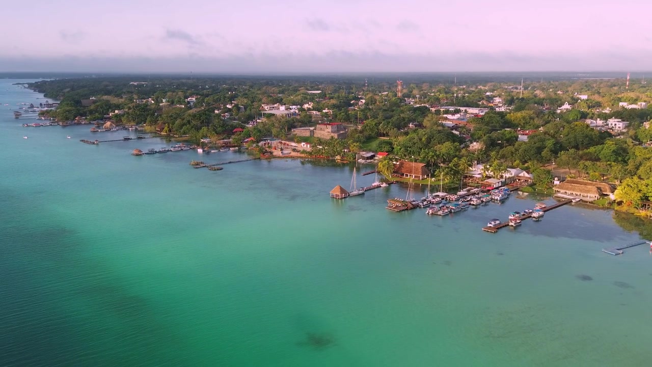 Ripresa con drone che avanza attraverso la laguna verso lo storico Forte di San Felipe all'alba, con barche a vela, porti turistici e case del villaggio in riva al lago lungo la costa turchese di Bacalar, Messico