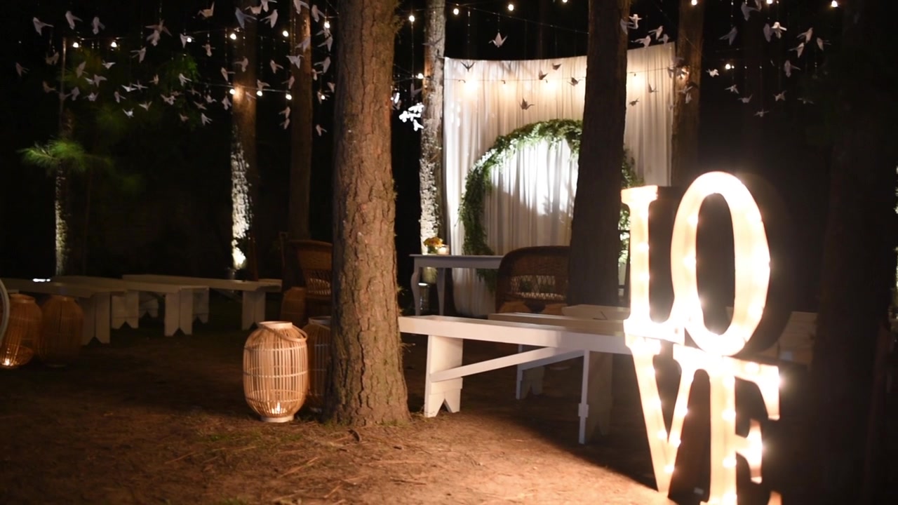Allestimenti per cerimonia nuziale notturna in stile vintage, un altare con lettere LOVE illuminate, sedie e luci romantiche sugli alberi, ripresa panoramica