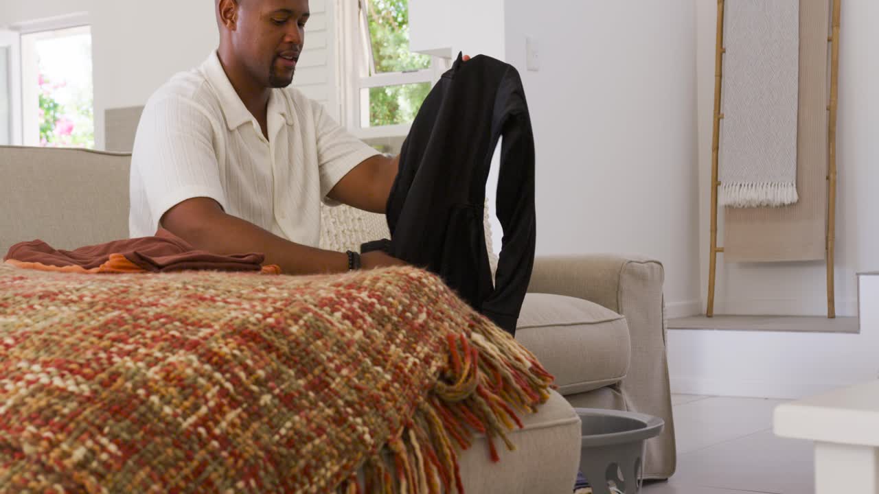 Adult African American man sitting on sofa, adjusting stack, taking dark pants, folding into basket