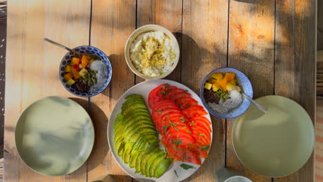 overhead view of a healthy breakfast table with sliced avocado, tomato, cottage cheese and chia pudding with fruit on a rustic wood surface