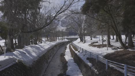 Evoking-a-sense-of-peacefulness,-the-camera-reveals-the-picturesque-scene-of-a-water-stream-peacefully-weaving-its-way-through-the-snow-covered-city-of-Hakuba-during-the-day