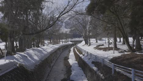 Die-Kamera-Weckt-Ein-Gefühl-Der-Ruhe-Und-Zeigt-Die-Malerische-Szene-Eines-Wasserstroms,-Der-Sich-Tagsüber-Friedlich-Durch-Die-Schneebedeckte-Stadt-Hakuba-Schlängelt