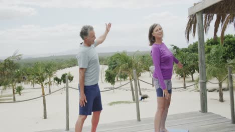 happy senior caucasian couple practicing yoga standing on beach sun deck, in slow motion