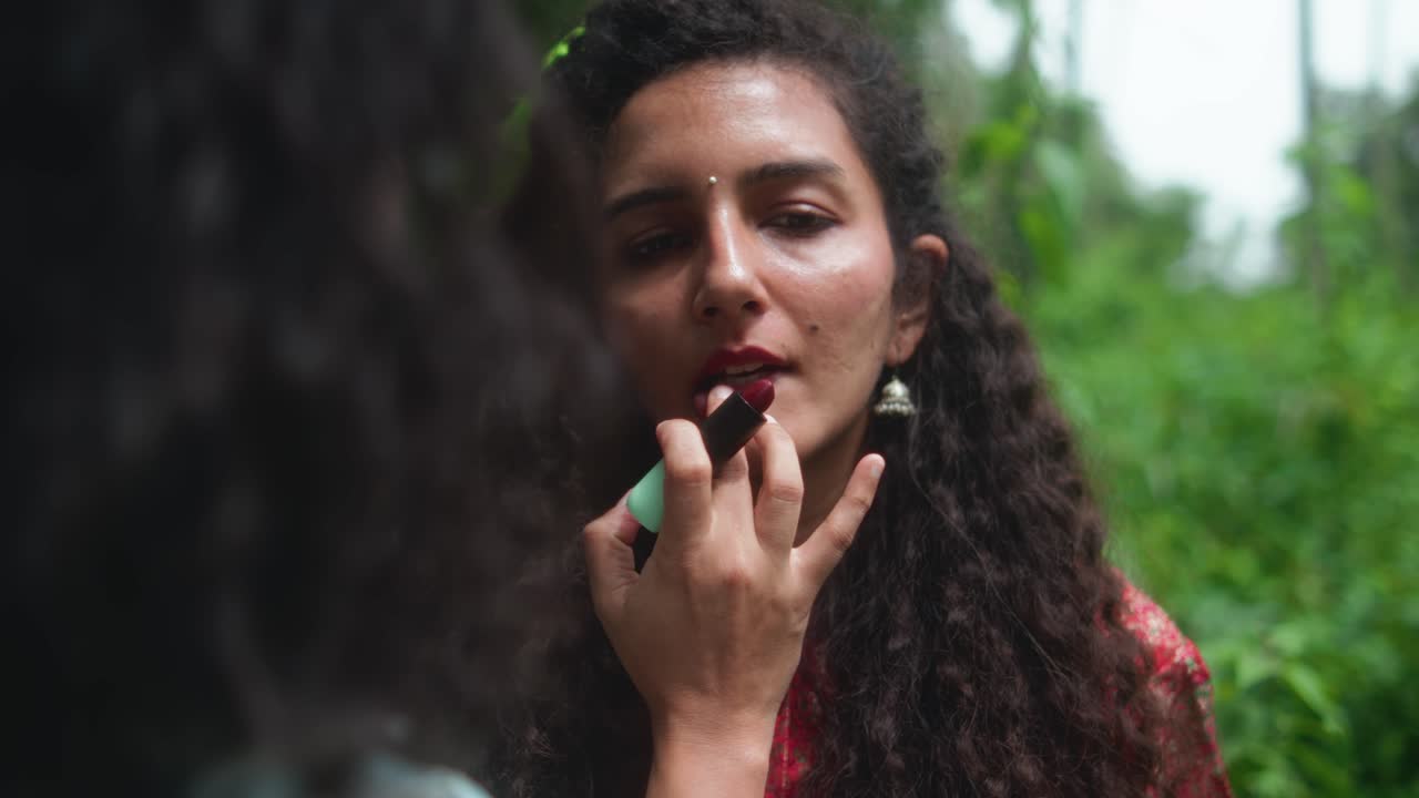 Close-up of a woman getting lipstick applied by a makeup artist. The makeup artist's head is in the foreground. The woman looks downward while lipstick is blended by hand